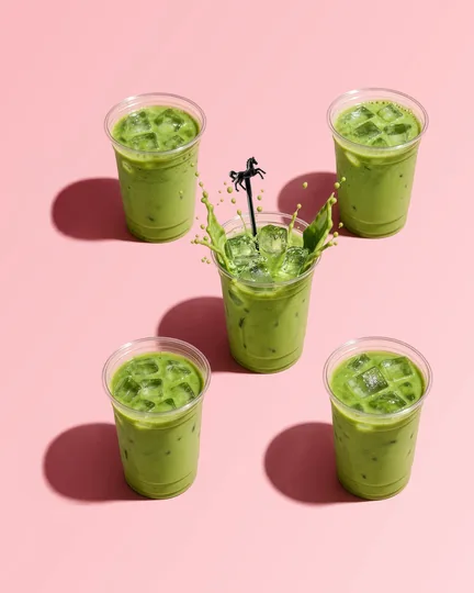 Iced matcha lattes on pink, a black horse stirrer splashing in the middle cup